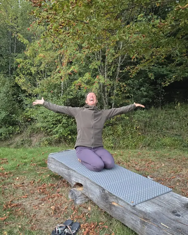 Christina kniet auf einem Baumstamm im Herbstwald und breitet die Arme aus