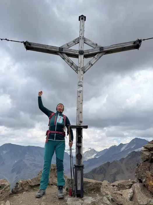 Christina steht jubelnd neben einem hölzernen Gipfelkreuz