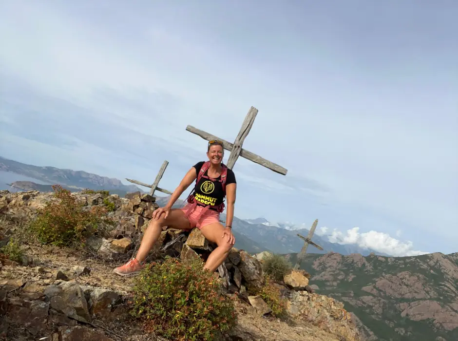 Christina sitzt lachend an einem Gipfelkreuz über weiten Bergketten