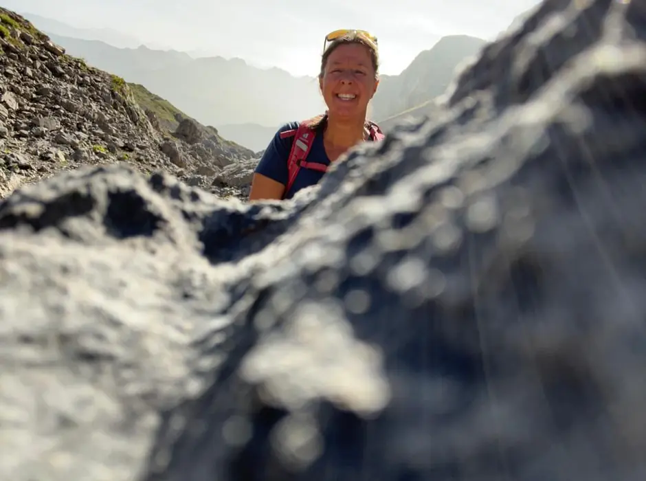 Christina lugt lachend hinter einem Felsen im Gebirge hervor